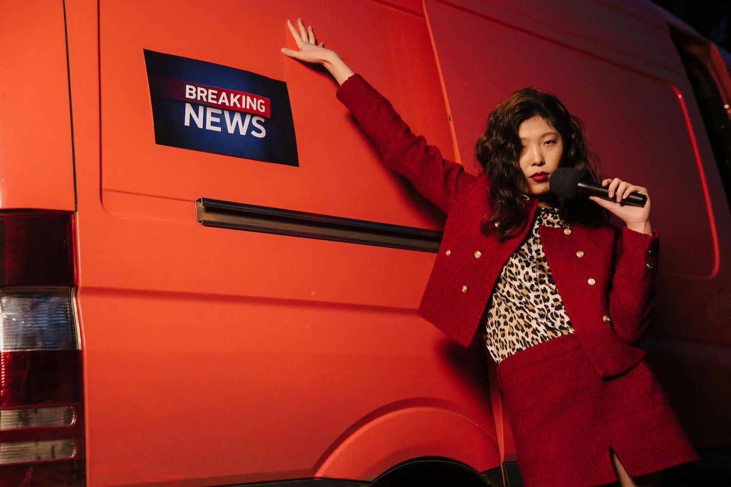 A woman with East Asian facial features stands by an orange van with her right hand's fingertips at the end of her outstretched right arm touching the side of the van. She wears a chic red suit with a leopard skin top and microphone in her left hand. With the sign 'Breaking News' on the van, she is a trendy news presenter.
