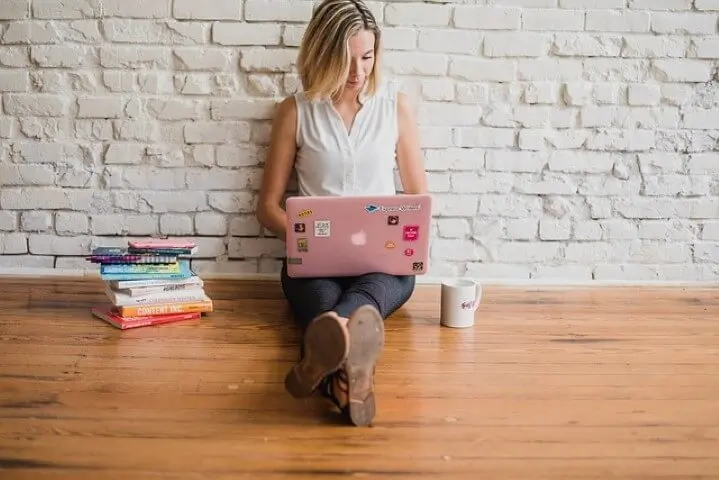 Woman sitting on a wooden floor against a white brick wall doing contnet marketing on a laptop