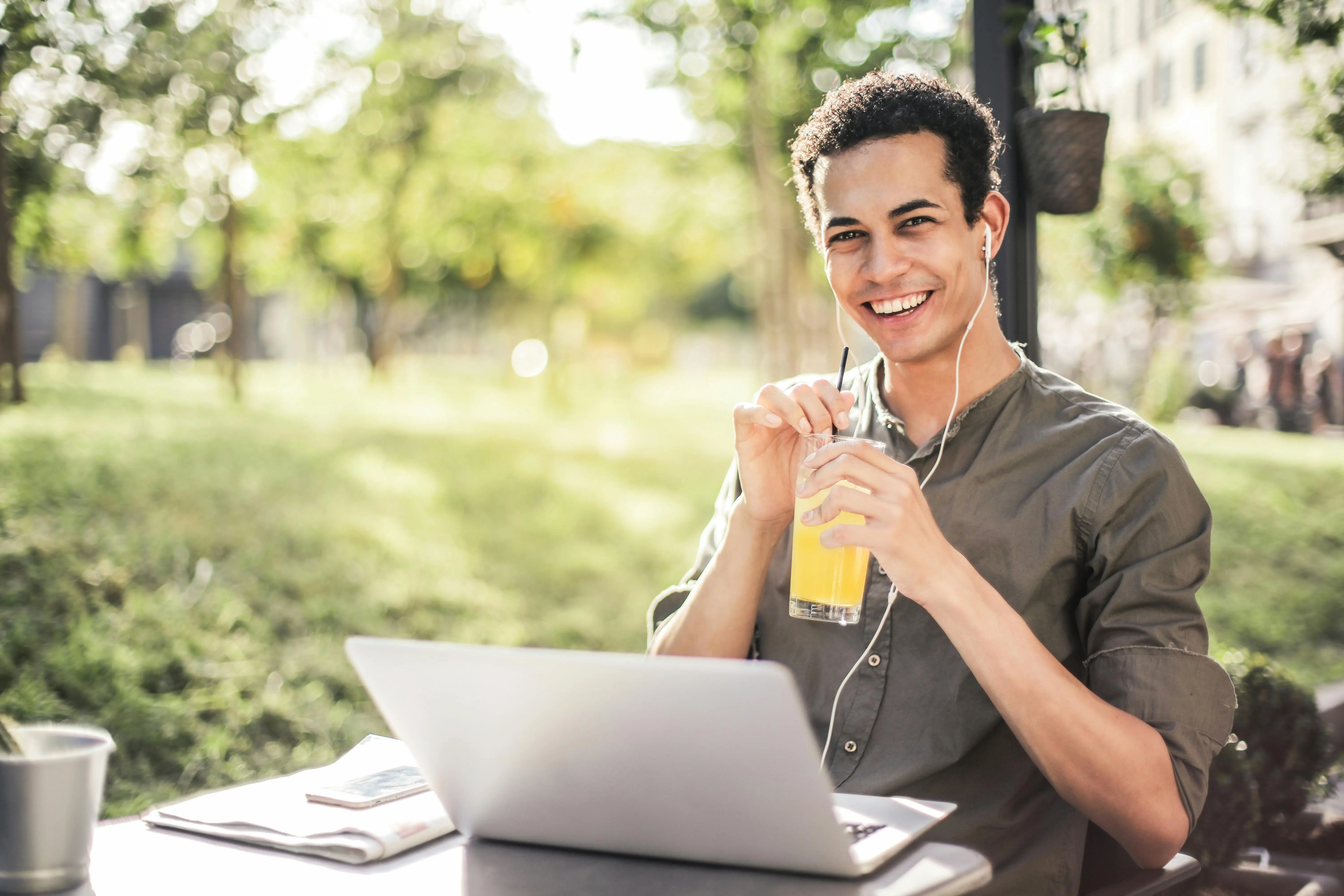 Happy man sitting at an outside table in a park. He has his laptop in front of him and an orange drink in his hand