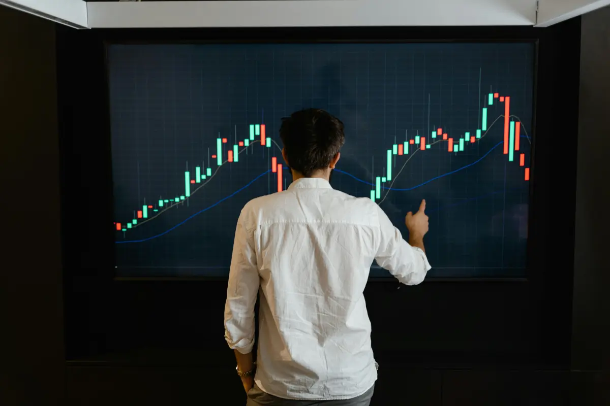 The back of a man standing in front of a screen. The screen shows the green and red bars of a candlestick chart