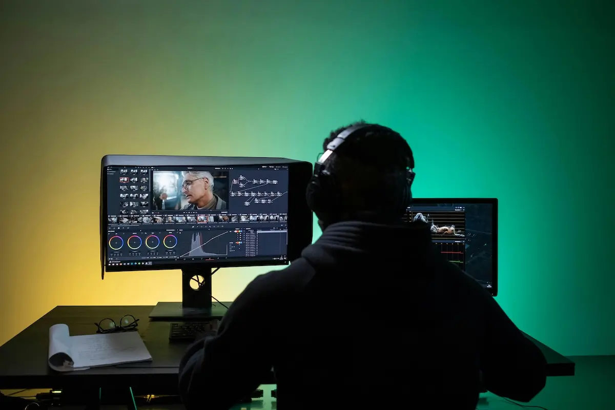 Back of a shadowed man at a desk. He's editing a video on an editing software. The wall behind the desk is lit up by a yellow light on the left and green light on the right