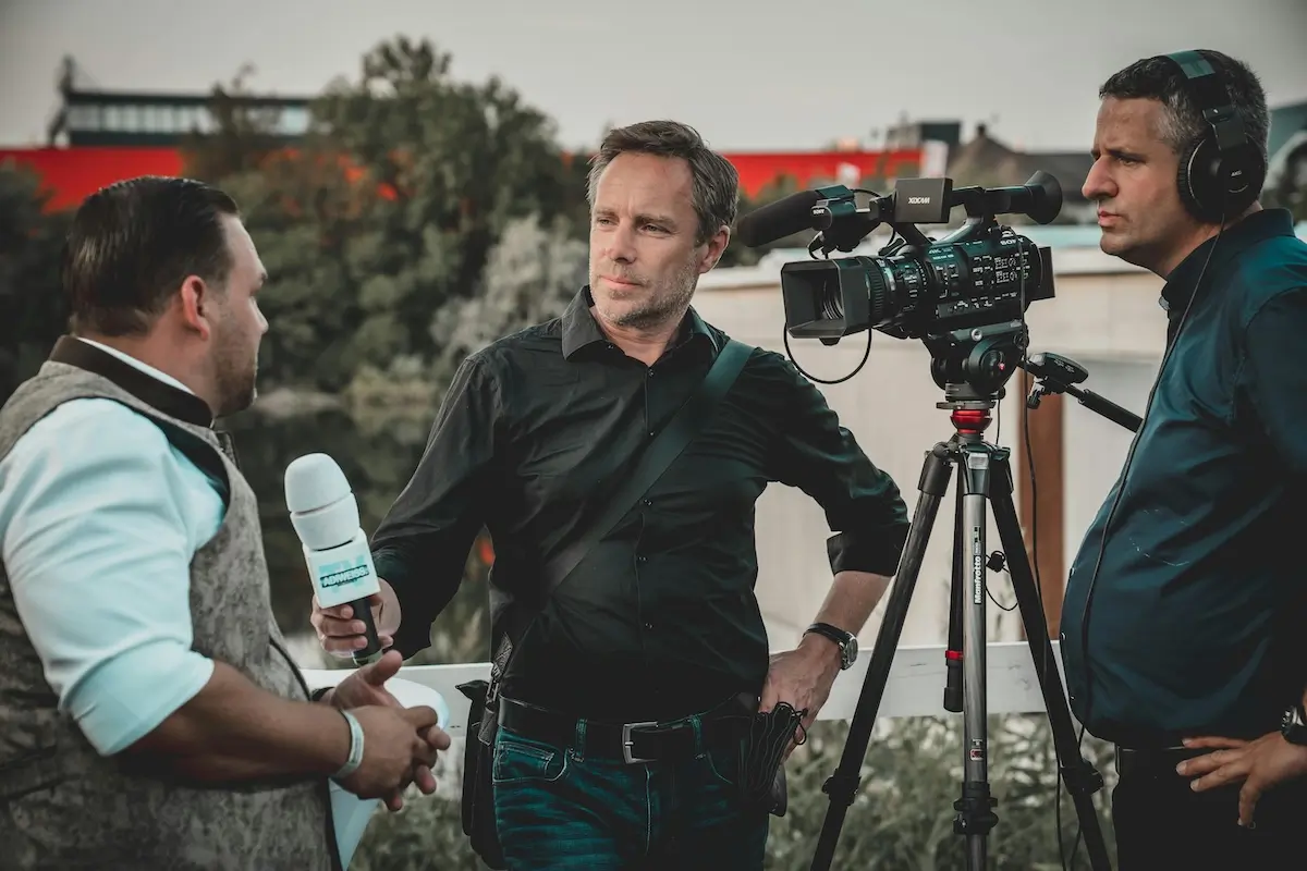 Male interviewing another make outside. Cameraman is filming it.