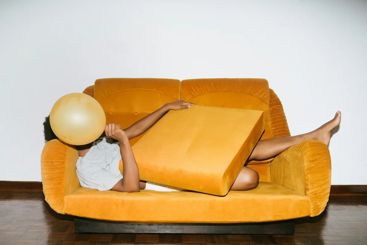 Person lying on an orange sofa covering their body with a cusion and face withan orange ballon