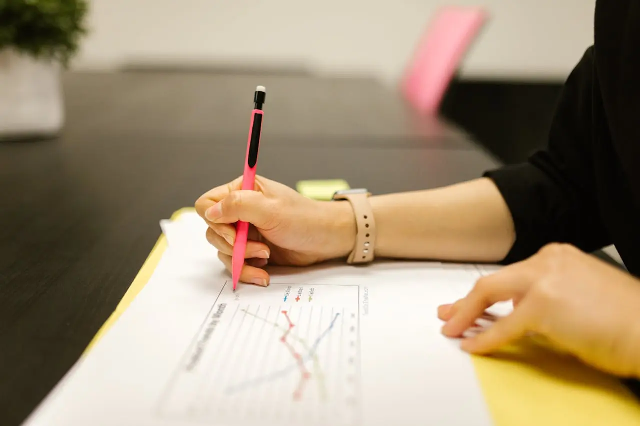 A female's hands on top of a piece of paper with the supply chain graph on it. She holds a pink pencil in her right hand. By RDNE on Pexels. com