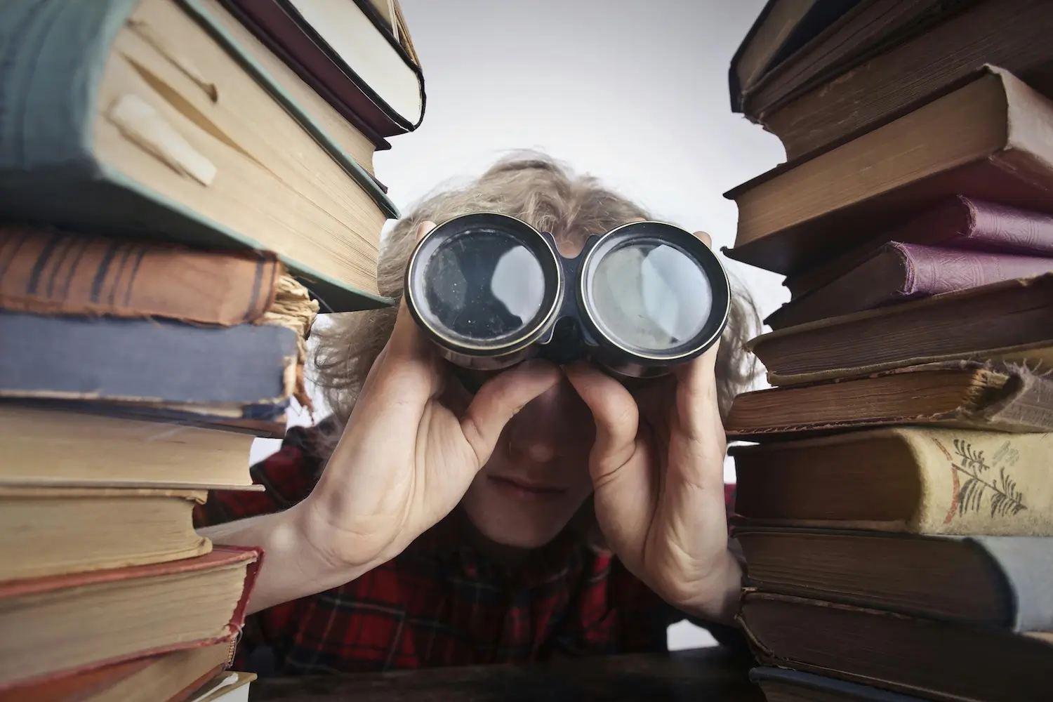 A person between piled books looking through binoculars at user-generated content by Andrea Piacquadio on Pexels. com