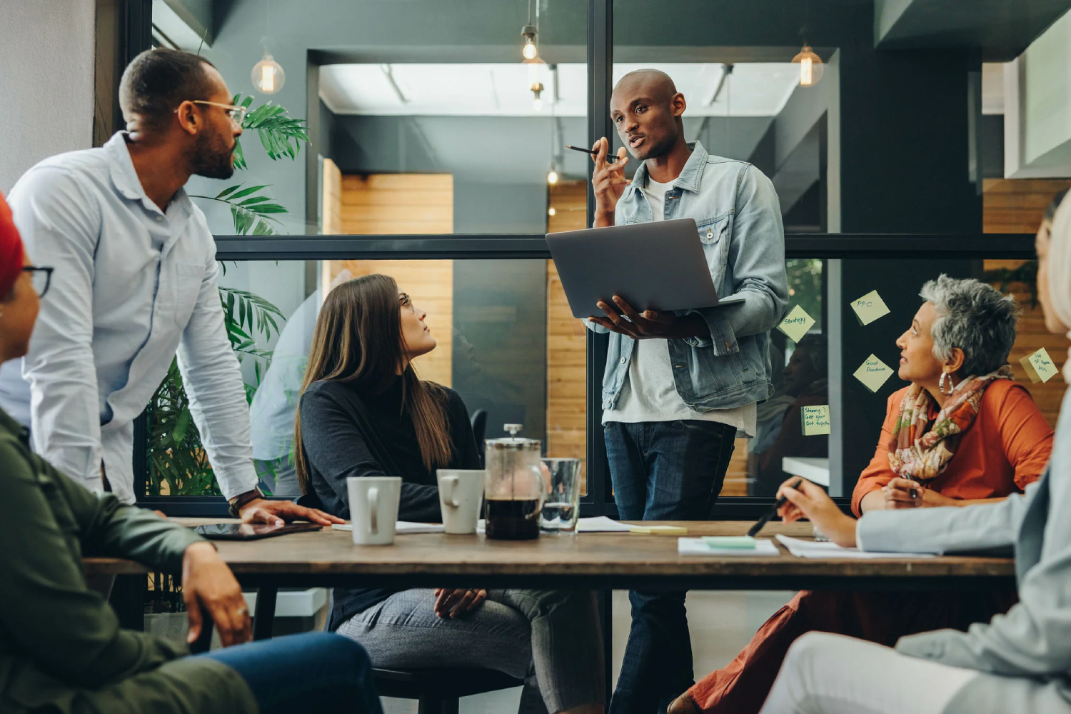 Five people have a meeting in a modern office. One person stands and speaks, holding a laptop, while the others sit at a table listening. Coffee cups and papers are on the table.