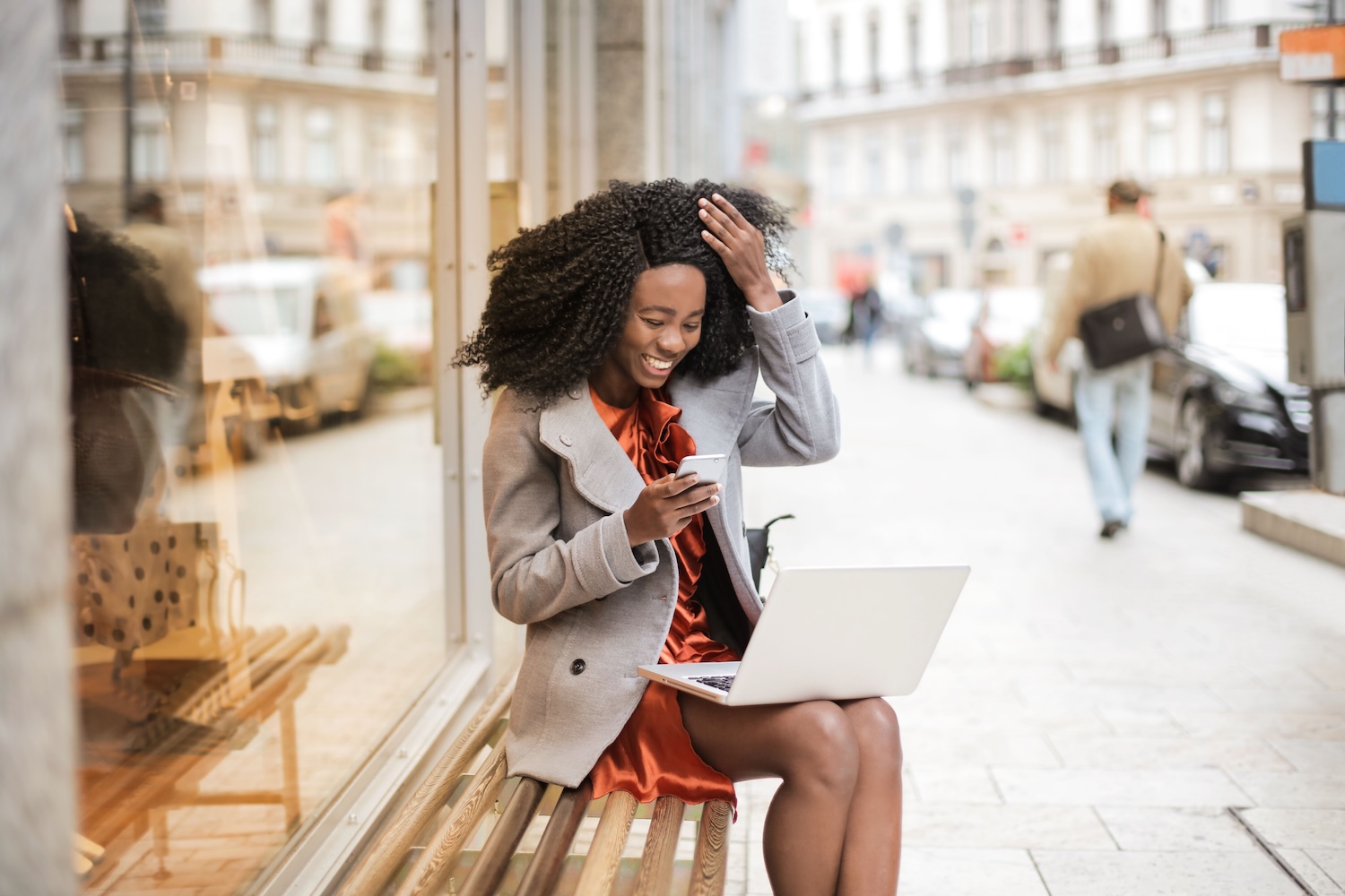 A young black woman sitting outside a shop with her laptop on her lap and a phone in her right hand. She is smiling by Andrea Piacquadio on Pexels